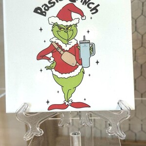 May include: A white ceramic tile with a green Grinch character wearing a red Santa hat and holding a blue cup. The tile has the text "Basic Grinch" in black lettering.