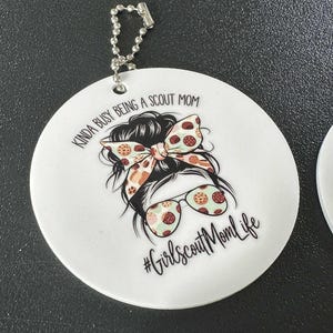 May include: White, round keychain with a graphic of a woman's hairstyle, sunglasses, and a bow. Text reads "Kinda busy being a Scout Mom" and "#GirlscoutMomLife". Includes a silver ball chain.