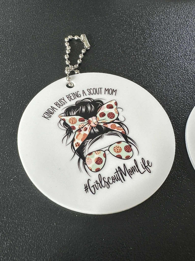 May include: White, round keychain with a graphic of a woman's hairstyle, sunglasses, and a bow. Text reads "Kinda busy being a Scout Mom" and "#GirlscoutMomLife". Includes a silver ball chain.