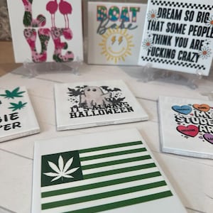 May include: A collection of white square ceramic tiles with various designs. One tile features the word "LOVE" with flamingo and floral patterns. Other tiles display phrases like "Boat Babe" and "Dream so big that some people think you are fucking crazy."