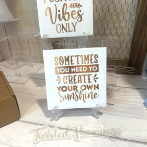 May include: Two white ceramic tiles with gold lettering on clear acrylic stands. The top tile reads "Positive Vibes Only" and the bottom tile reads "Sometimes you need to create your own sunshine". The tiles are on a white wooden surface with a clear acrylic sign that reads "Twisted Flamingos".