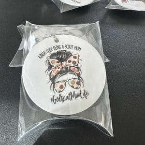 May include: White round ornament with a graphic of a woman with a messy bun, sunglasses, and a patterned bow. Text reads "Kinda busy being a Scout Mom" and "#GirlscoutMomLife". Packaged in a clear plastic sleeve.