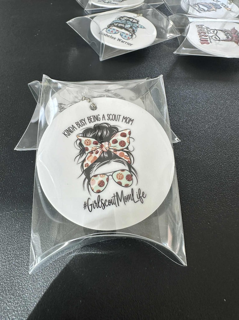May include: White round ornament with a graphic of a woman with a messy bun, sunglasses, and a patterned bow. Text reads "Kinda busy being a Scout Mom" and "#GirlscoutMomLife". Packaged in a clear plastic sleeve.