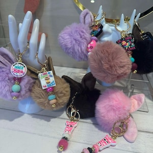 May include: A collection of keychains with fluffy pom-poms and decorative beads. Some keychains feature bunny ears, while others have charms with text like "Inner Bitch" and "I take my coffee on the rocks". The keychains are in various colors.