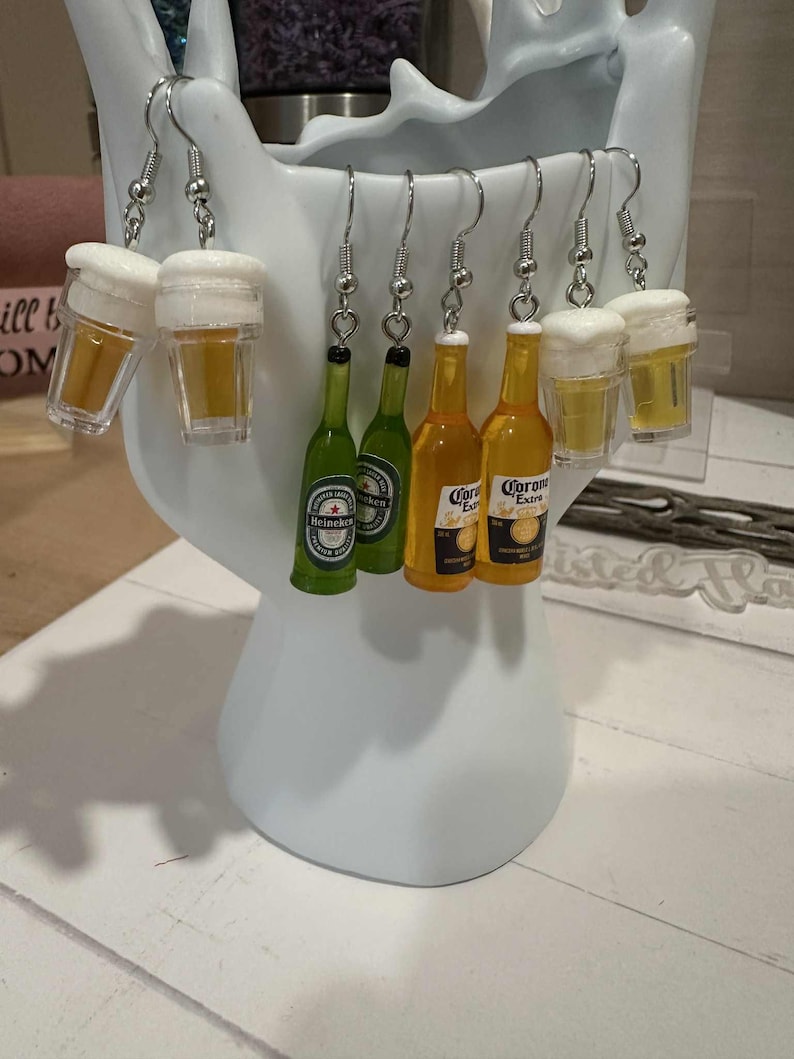 May include: A collection of novelty earrings featuring miniature beer glasses and beer bottles. The earrings include clear plastic beer glasses with white foam tops, and green Heineken and yellow Corona beer bottle designs.