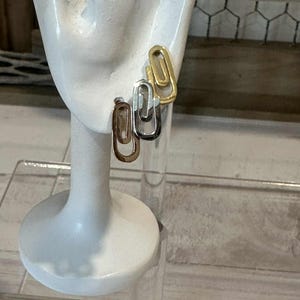 May include: Three pairs of paperclip-shaped earrings in gold, silver, and bronze tones are displayed on a white ear-shaped jewelry holder. The earrings are simple and modern, with a polished finish. The jewelry holder is on a clear acrylic stand.