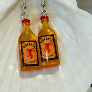 May include: A pair of novelty earrings featuring miniature Fireball whiskey bottles. The bottles are orange with red caps and black and orange labels. The earrings hang from silver-colored fish hook ear wires.
