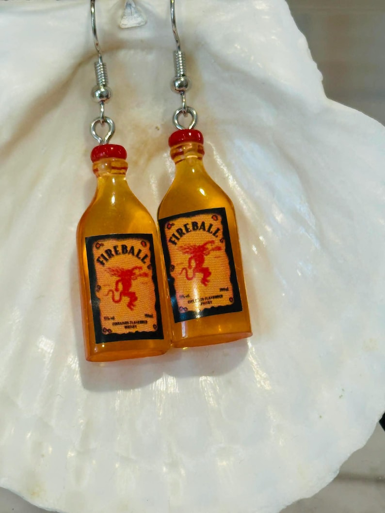 Puede incluir: Un par de pendientes originales con botellas de whisky Fireball en miniatura. Las botellas son de color naranja con tapones rojos y etiquetas negras y naranjas. Cuelgan de ganchos para las orejas de color plateado.