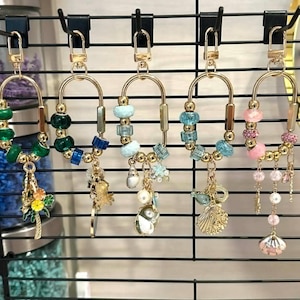 May include: A collection of gold-toned keychains with various charms and colorful beads. Each keychain features a unique design, including floral, sea-themed, and abstract elements. The keychains are displayed on a black wire grid.