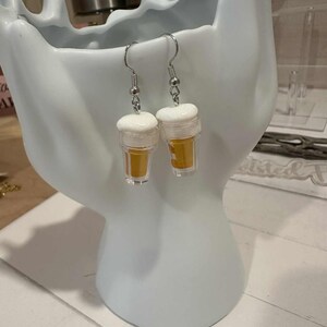 May include: A pair of novelty earrings shaped like miniature beer glasses. Each earring features a clear plastic cup filled with a golden liquid, topped with a white foam. The earrings hang from silver-colored fish hook ear wires.