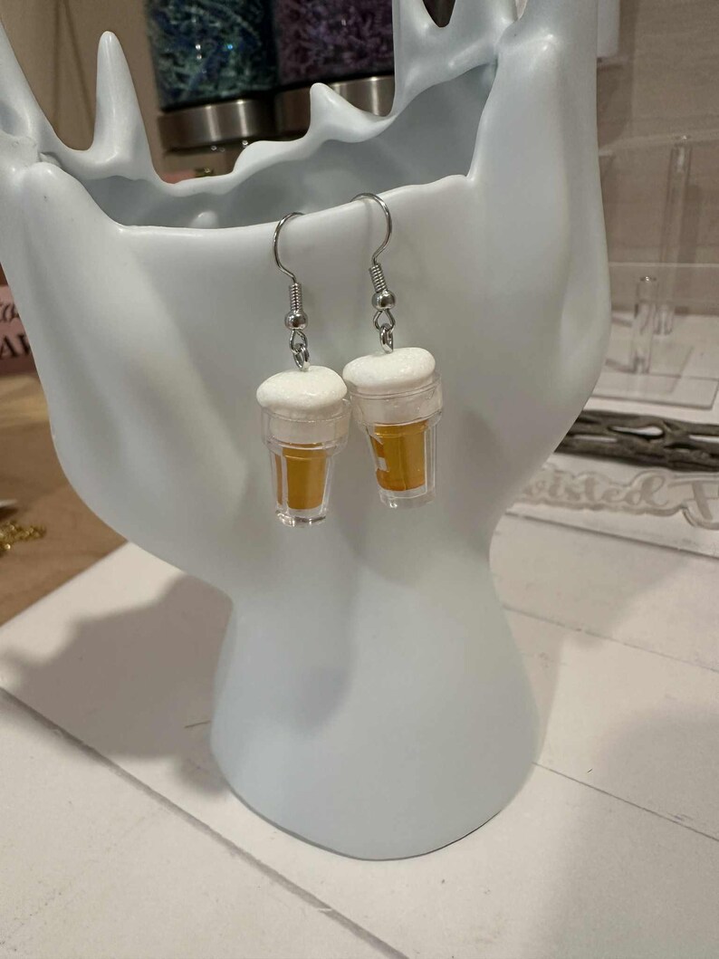 May include: A pair of novelty earrings shaped like miniature beer glasses. Each earring features a clear plastic cup filled with a golden liquid, topped with a white foam. The earrings hang from silver-colored fish hook ear wires.