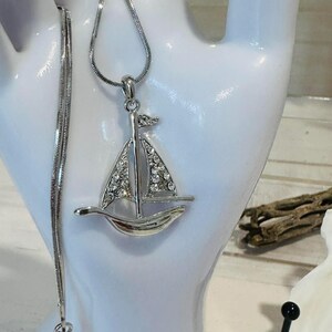 May include: A silver-toned sailboat pendant necklace. The sailboat features a sail encrusted with clear crystals. The necklace chain is a delicate snake chain. The pendant is displayed on a white surface.