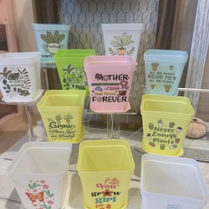 Puede incluir: Surtido de macetas cuadradas de colores con diferentes lemas y diseños. Las macetas presentan lemas como "Plant Mama", "Grow Where You Are Planted", "Mother, Plant the Seeds of Love That Bloom Forever", "Never Enough Plants", "You Grow Girl" y "Crazy Plant Lady".