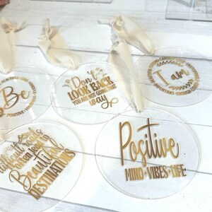 May include: Clear acrylic coasters with gold lettering featuring inspirational quotes like "Be Positive" and "Difficult roads often lead to beautiful destinations." Each coaster has a ribbon attached.