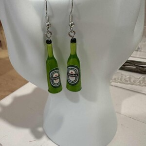 May include: A pair of green Heineken beer bottle earrings with silver-colored hooks. The earrings feature miniature bottle replicas with the Heineken logo and a black bottle cap.