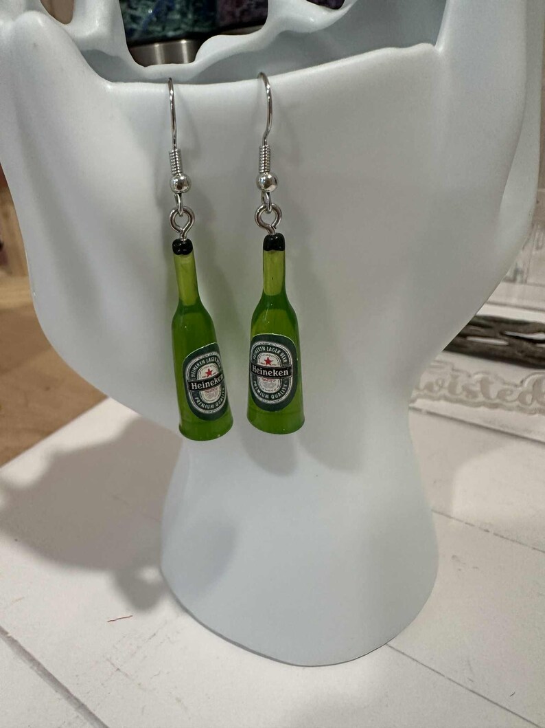 May include: A pair of green Heineken beer bottle earrings with silver-colored hooks. The earrings feature miniature bottle replicas with the Heineken logo and a black bottle cap.