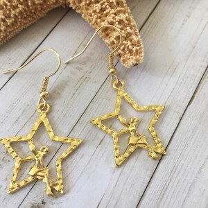 May include: Gold-colored star-shaped earrings with a textured finish. Each earring features a cutout design of a figure inside the star. The earrings hang from gold-colored fish hook ear wires. The earrings are displayed on a light wood surface.