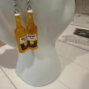 May include: A pair of dangle earrings featuring miniature Corona Extra beer bottles. The bottles are yellow with white and black labels, and white bottle caps. The earrings hang from silver-colored fish hook ear wires.
