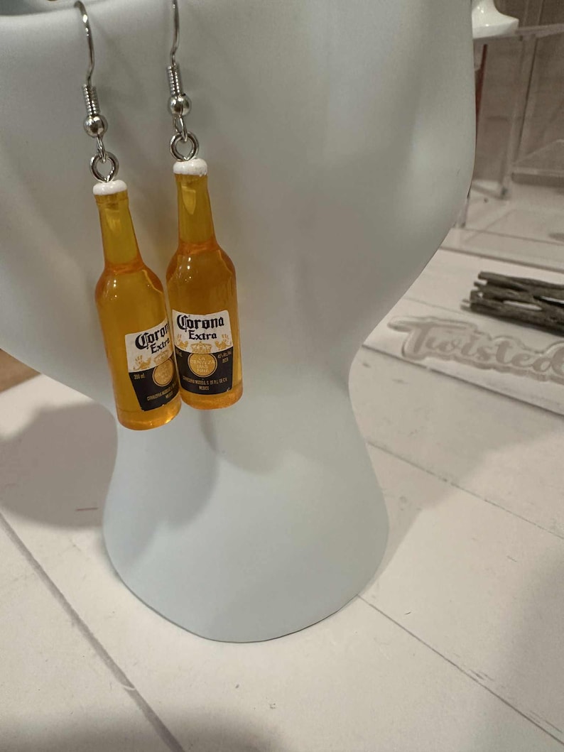 May include: A pair of dangle earrings featuring miniature Corona Extra beer bottles. The bottles are yellow with white and black labels, and white bottle caps. The earrings hang from silver-colored fish hook ear wires.