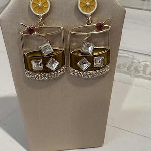 May include: A pair of gold-toned cocktail-themed earrings. Each earring features a glass design with ice cubes, a cherry, and a lemon slice. The base of the glass is embellished with clear crystals. The earrings are displayed on a beige stand.