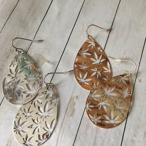 May include: A pair of dangle earrings with a teardrop shape. One pair is silver with a leaf pattern and the other pair is gold with a leaf pattern.