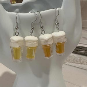 May include: Four beer-themed earrings. Each earring features a miniature clear plastic cup filled with a yellow liquid, topped with white foam. The earrings hang from silver-colored fish hook ear wires.