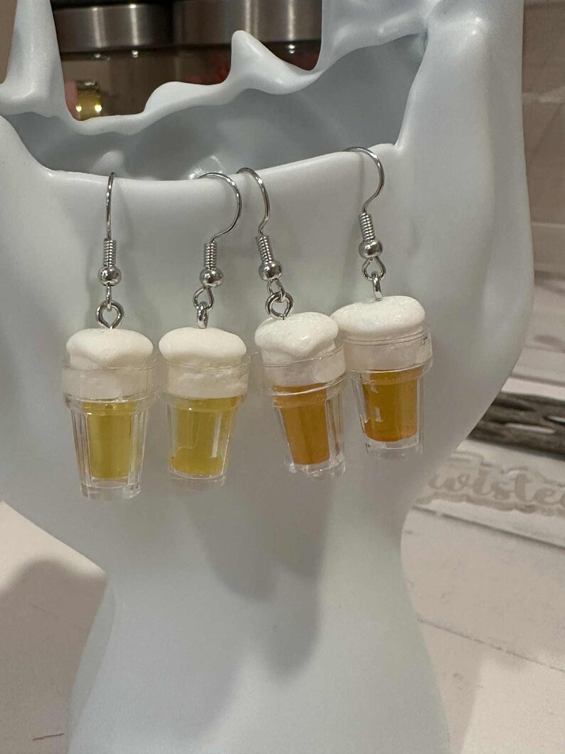May include: Four beer-themed earrings. Each earring features a miniature clear plastic cup filled with a yellow liquid, topped with white foam. The earrings hang from silver-colored fish hook ear wires.