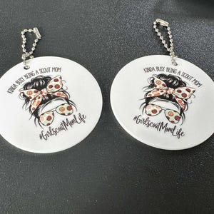 May include: Two white round keychains with a printed design. The design features a woman's face with sunglasses and a bow, and the text "KINDA BUSY BEING A SCOUT MOM" and "#GirlscoutMomLife". Each keychain has a silver chain.