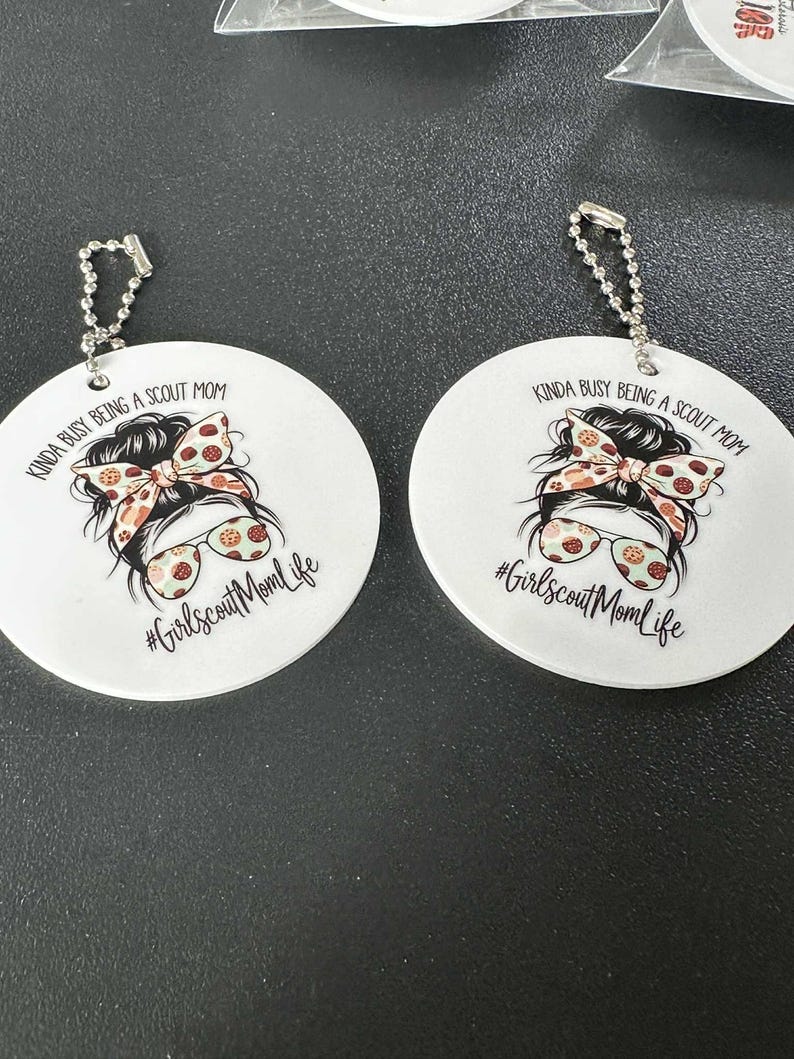 May include: Two white round keychains with a printed design. The design features a woman's face with sunglasses and a bow, and the text "KINDA BUSY BEING A SCOUT MOM" and "#GirlscoutMomLife". Each keychain has a silver chain.