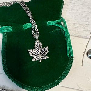 May include: A silver-colored necklace with a maple leaf pendant rests on a dark green velvet pouch. The necklace chain is visible, and the pouch has a green drawstring closure. The background is a light-colored wooden surface.