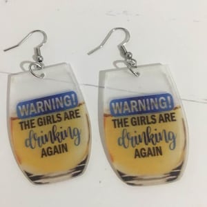 May include: A pair of novelty earrings with a clear, glass-shaped design. Each earring features a blue rectangle with the word "WARNING!" above the text "THE GIRLS ARE drinking AGAIN" in blue script. The earrings have silver-colored fish hook closures.