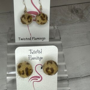 May include: A pair of leopard print pom-pom earrings on a white card with the text "Twisted Flamingo".