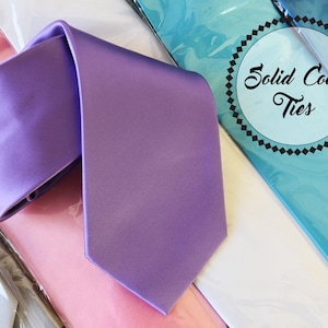 Può includere: Una collezione di cravatte tinta unita in vari colori, tra cui lavanda, rosa, grigio e blu. Le cravatte sono esposte su uno sfondo bianco con un cartello che recita "Cravatte tinta unita".