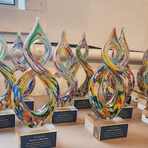 May include: A collection of flame-shaped glass awards featuring a multi-colored swirl pattern of red, yellow, blue, and green. Each award is mounted on a clear rectangular base with a blue plaque inscribed with the text To honor your Service to Education.