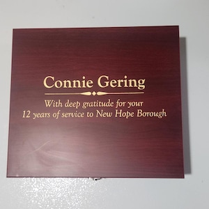 May include: A rectangular wooden plaque with a dark reddish-brown finish. The text "Connie Gering" is displayed in gold lettering, along with a message of gratitude for 12 years of service to New Hope Borough.