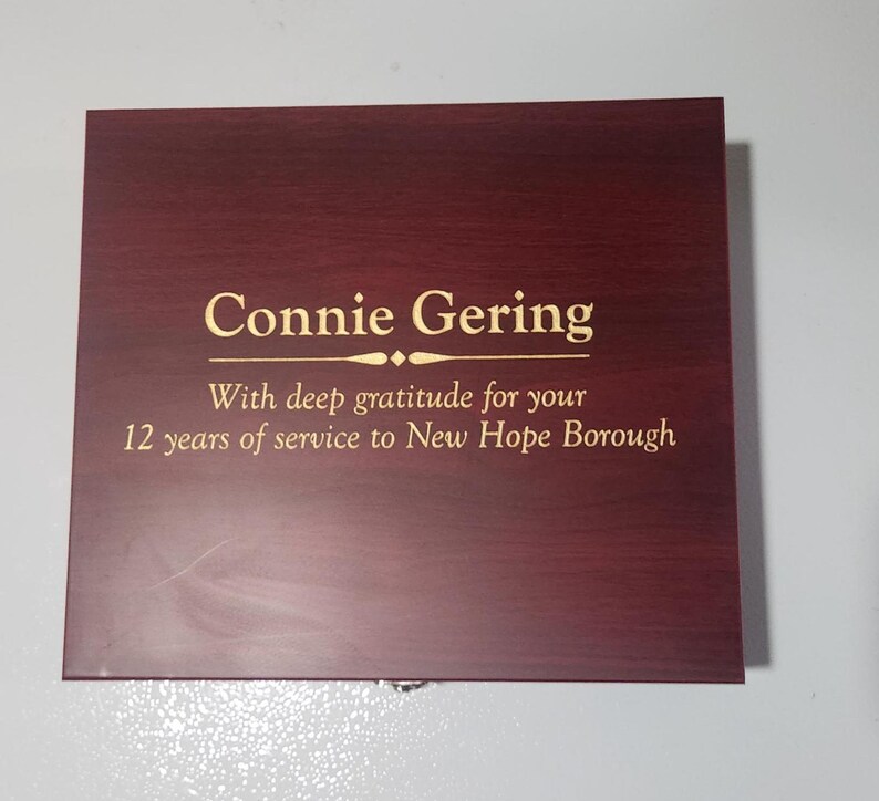 May include: A rectangular wooden plaque with a dark reddish-brown finish. The text "Connie Gering" is displayed in gold lettering, along with a message of gratitude for 12 years of service to New Hope Borough.