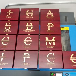 Collection of personalized groomsmen gift boxes with engraved gold initials, names, roles like groomsman, best man, usher, and wedding date 09.28.2019. Each rosewood-finished box features custom laser engraving, perfect for wedding party gifts. Ideal for groomsmen proposals, bachelor parties, or keepsakes. Displayed on a worktable, showcasing bulk order customization for a wedding event.