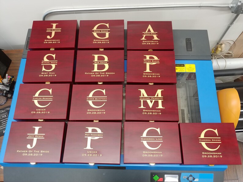 Collection of personalized groomsmen gift boxes with engraved gold initials, names, roles like groomsman, best man, usher, and wedding date 09.28.2019. Each rosewood-finished box features custom laser engraving, perfect for wedding party gifts. Ideal for groomsmen proposals, bachelor parties, or keepsakes. Displayed on a worktable, showcasing bulk order customization for a wedding event.