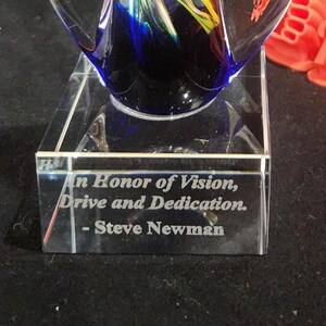 May include: A clear glass award featuring a deep blue and yellow swirl design at the top. The rectangular base is engraved with the text: In Honor of Vision, Drive and Dedication. - Steve Newman. The object is made of polished glass.