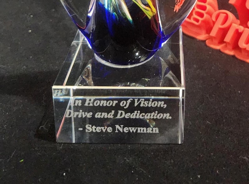 May include: A clear glass award featuring a deep blue and yellow swirl design at the top. The rectangular base is engraved with the text: In Honor of Vision, Drive and Dedication. - Steve Newman. The object is made of polished glass.