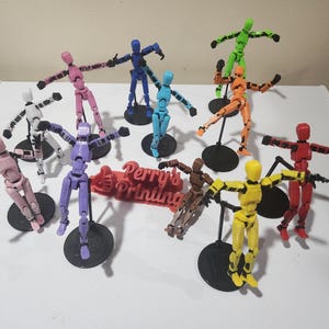 May include: A collection of articulated action figures in various colors, including pink, blue, green, and yellow. Each figure is mounted on a black base and has movable joints. A red sign with the text "Perry's Printing" is also present.