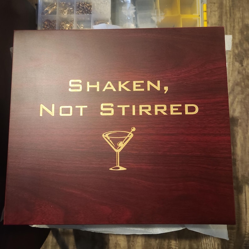 Elegant rosewood cocktail gift box with engraved gold text reading 'SHAKEN, NOT STIRRED' and featuring a martini glass illustration with an olive. The polished wood finish and laser-engraved design make it a perfect gift for mixology enthusiasts or James Bond fans. Ideal for barware sets or groomsmen gifts.