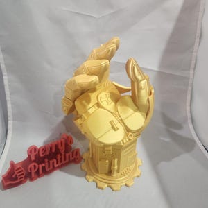 May include: A gold-colored, 3D-printed robotic hand sculpture. The hand is mounted on a detailed base with gear-like elements. A red, thumb-up shaped sign with the text "Perry's Printing" is also present.
