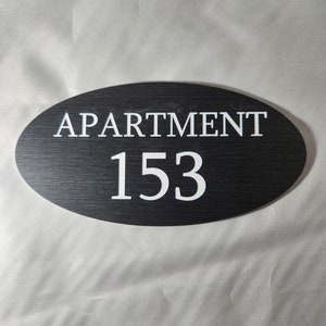 May include: Black oval sign with white text that reads "APARTMENT 153".