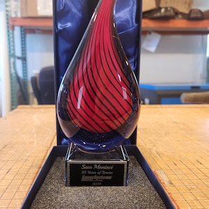 May include: A teardrop-shaped glass award featuring a red and black swirl pattern, mounted on a clear glass base. The base displays the text: Sam Moniaci, 25 Years of Service, SaverSystems, 2024. The award is presented in a blue-lined display box.
