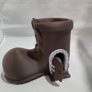 May include: A brown boot-shaped planter with a small door and arched doorway. The boot has laces and a textured surface. The door is open, revealing a dark interior. The planter is designed to hold plants or flowers.