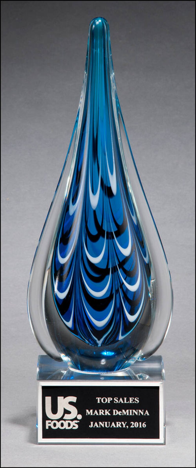 A blue glass award with white and black swirling designs on a clear glass base. The base has a black plaque with silver lettering that reads 'US Foods, Top Sales, Mark Deminna, January 2016.'