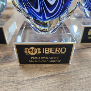 Close-up of a stunning blue and white hand-blown glass teardrop award with intricate swirl patterns, mounted on a clear rectangular base. The engraved plaque reads "IBERO American Action League, Inc. – President’s Award – Maria Esther Sanchez." Ideal for personalized corporate recognition, employee appreciation, or commemorative gifts. Elegant and artistic, perfect for display in any office or award showcase.