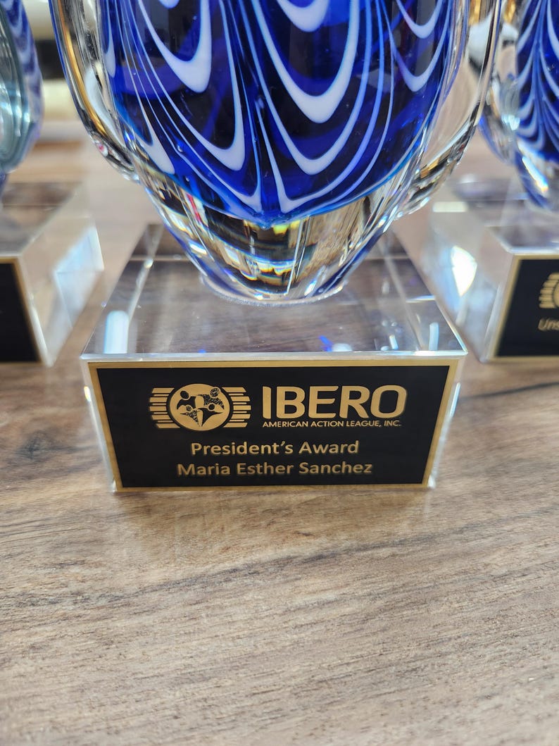 Close-up of a stunning blue and white hand-blown glass teardrop award with intricate swirl patterns, mounted on a clear rectangular base. The engraved plaque reads "IBERO American Action League, Inc. – President’s Award – Maria Esther Sanchez." Ideal for personalized corporate recognition, employee appreciation, or commemorative gifts. Elegant and artistic, perfect for display in any office or award showcase.