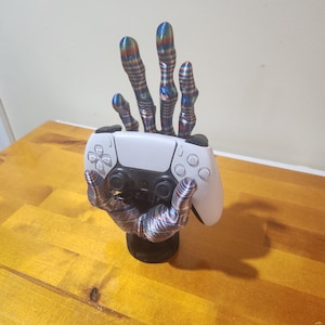May include: A 3D printed hand with a colorful striped pattern holds a white video game controller. The hand is on a black base.